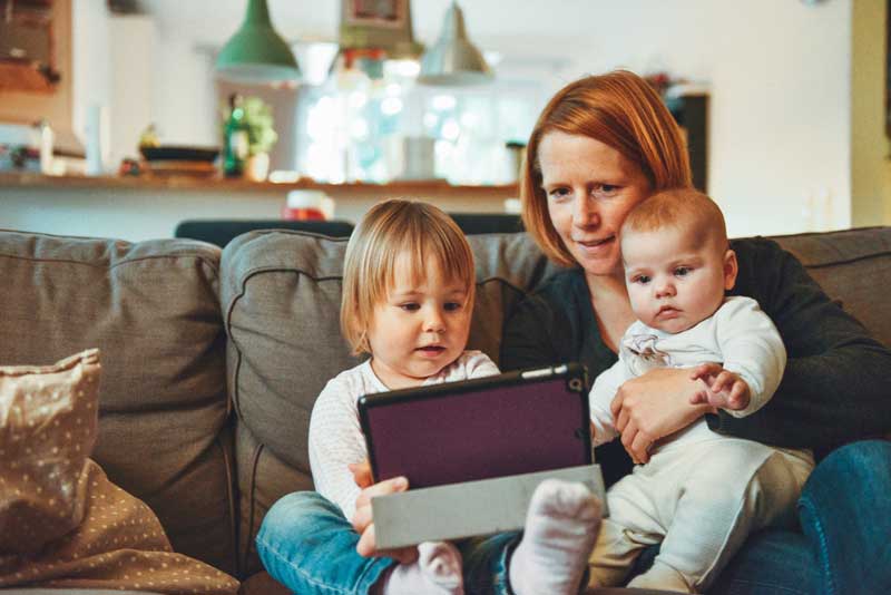 Mom sitting on the sofa with toddler next to her and baby in her lap looking at tablet computer.